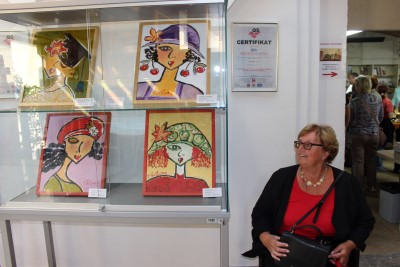 Tinka Šetina sedi ob stekleni vitrini, v kateri so štirje njeni živobarvni stilizirani ženski portreti.
